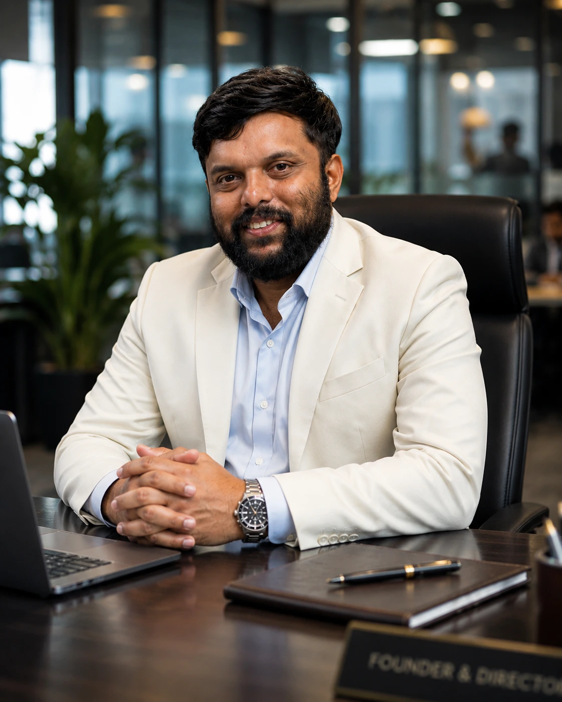 A professional South Indian businessman wearing an ivory-white suit and light blue shirt sits confidently in a modern office environment. The background features blurred workstations, employees, glass partitions, and indoor plants, creating a sophisticated corporate atmosphere. The subject maintains direct eye contact with the camera, conveying leadership, confidence, and professionalism.